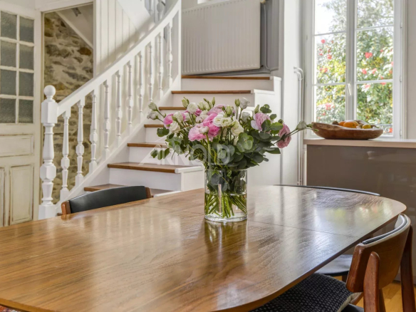 Wooden dining table with a glass vase of pink and white flowers and greenery, set in a bright room with a white staircase and large window in the background.