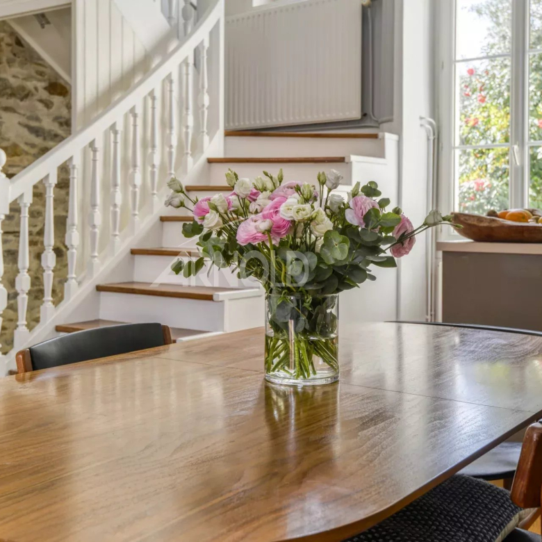 Wooden dining table with a glass vase of pink and white flowers and greenery, set in a bright room with a white staircase and large window in the background.