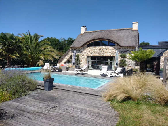 Stone house with thatched roof beside a long rectangular pool and wooden deck, surrounded by lounge chairs and tropical plants under a clear blue sky.