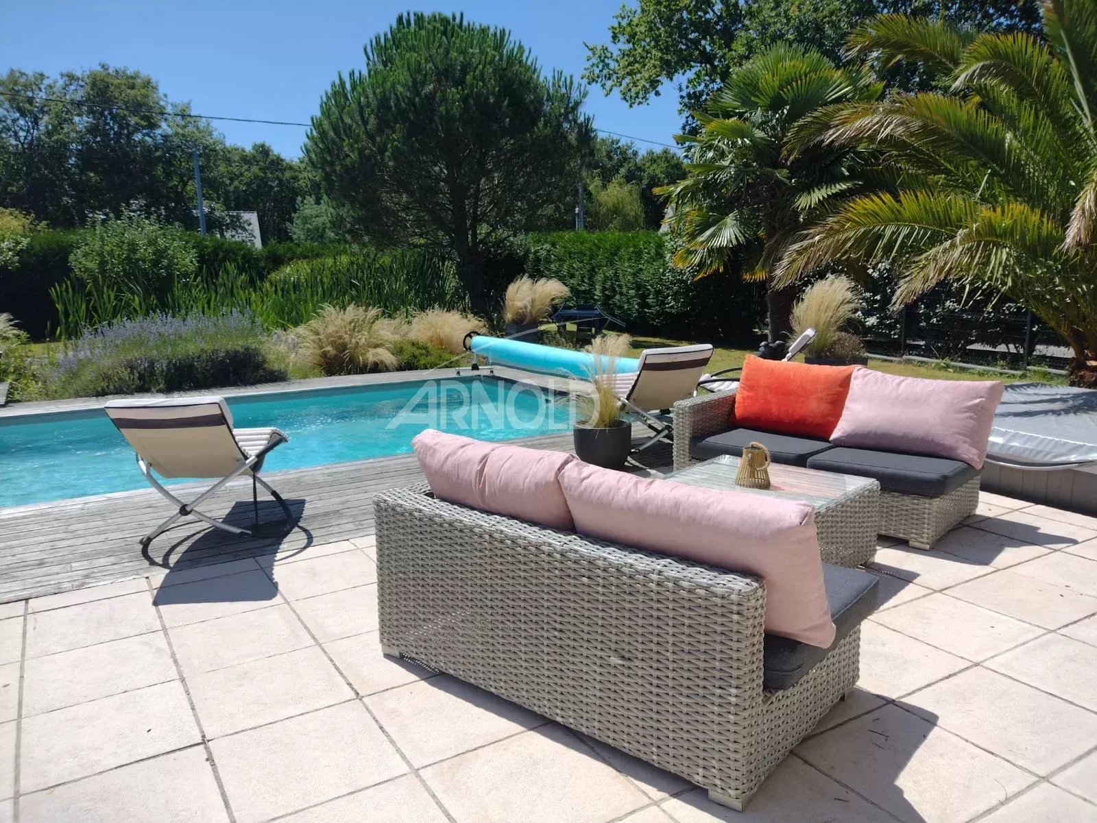 Outdoor poolside seating area with woven rattan sofa and chairs, pink cushions, a small table, and a bright blue pool in the background.