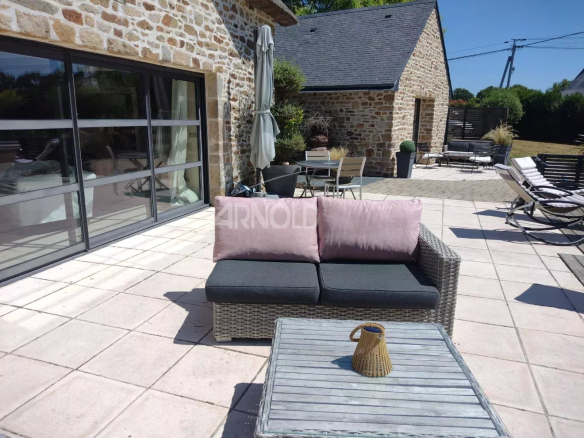 Sunny brick-and-stone patio with a wicker sofa and pink cushions facing a glass sliding-door building, plus lounge chairs and a coffee table on a tiled patio.
