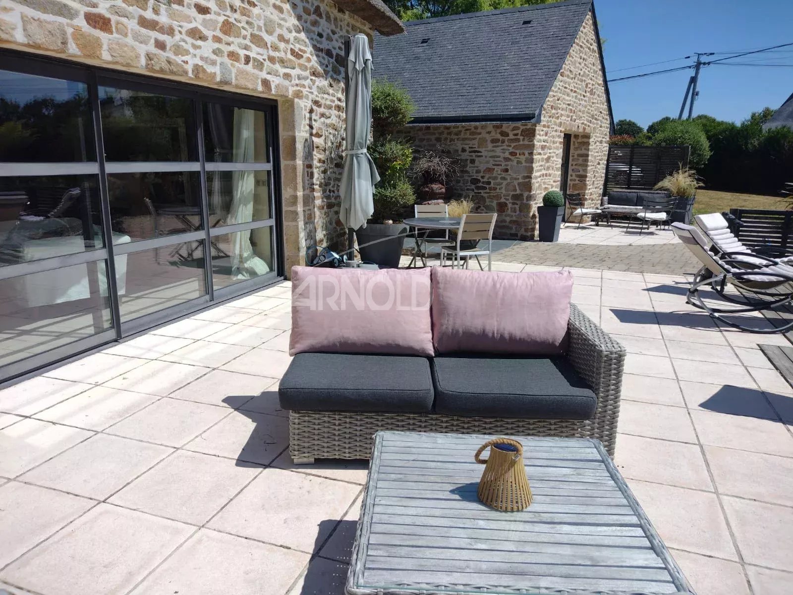 Sunny brick-and-stone patio with a wicker sofa and pink cushions facing a glass sliding-door building, plus lounge chairs and a coffee table on a tiled patio.