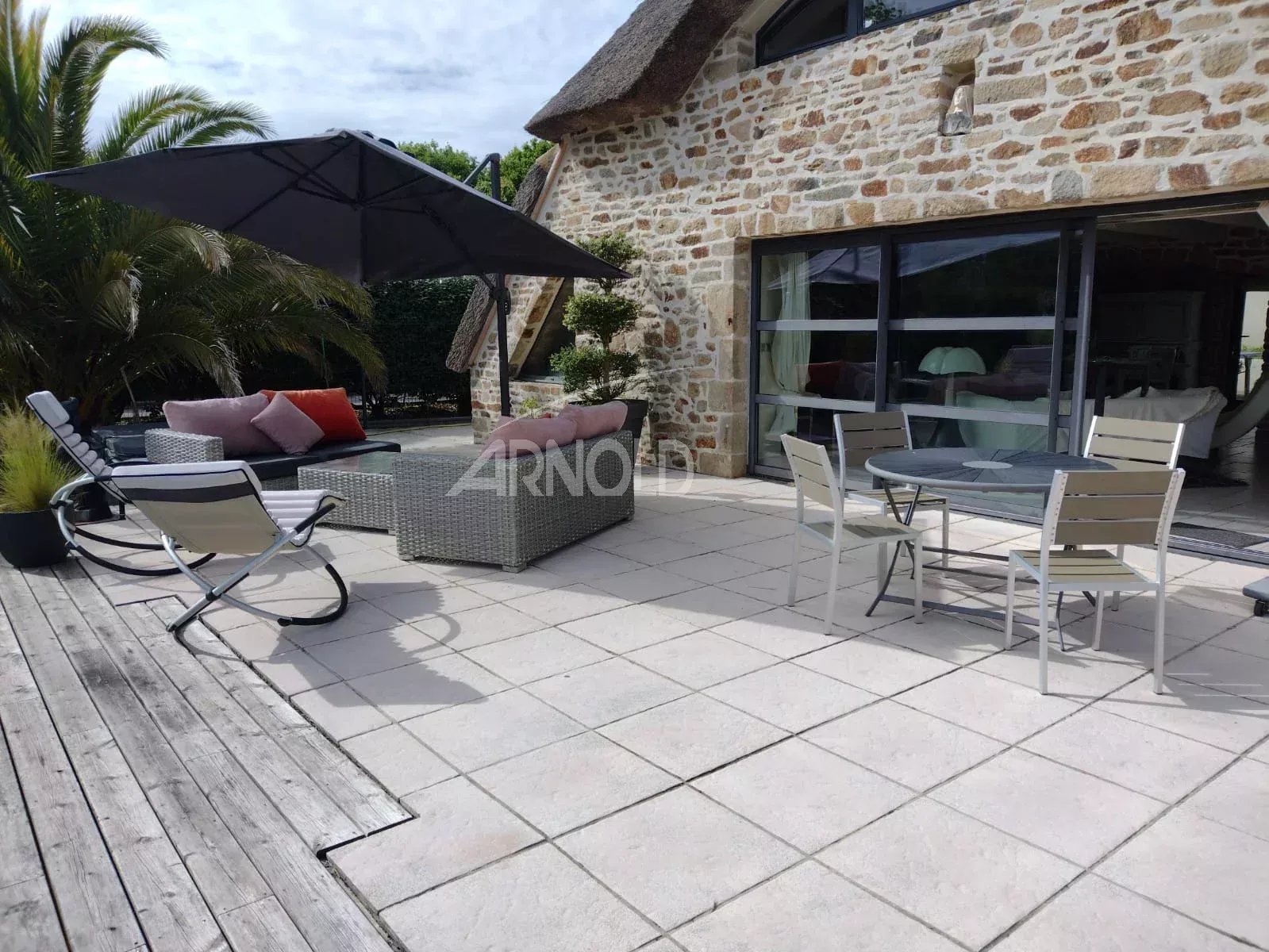 Outdoor patio with stone house, wicker sofa, cushioned seating, and a large dark umbrella on a tiled deck