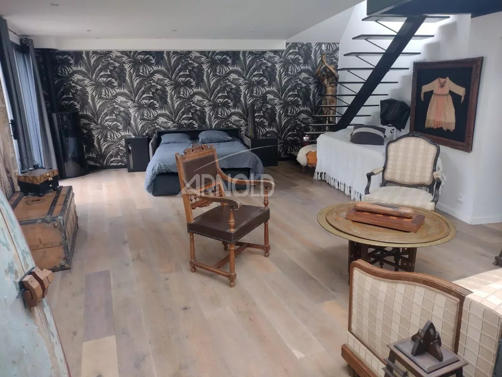 Loft living space with palm-patterned accent wall, a bed in the back, and vintage chairs around a round coffee table under a metal staircase, airy stone floor.
