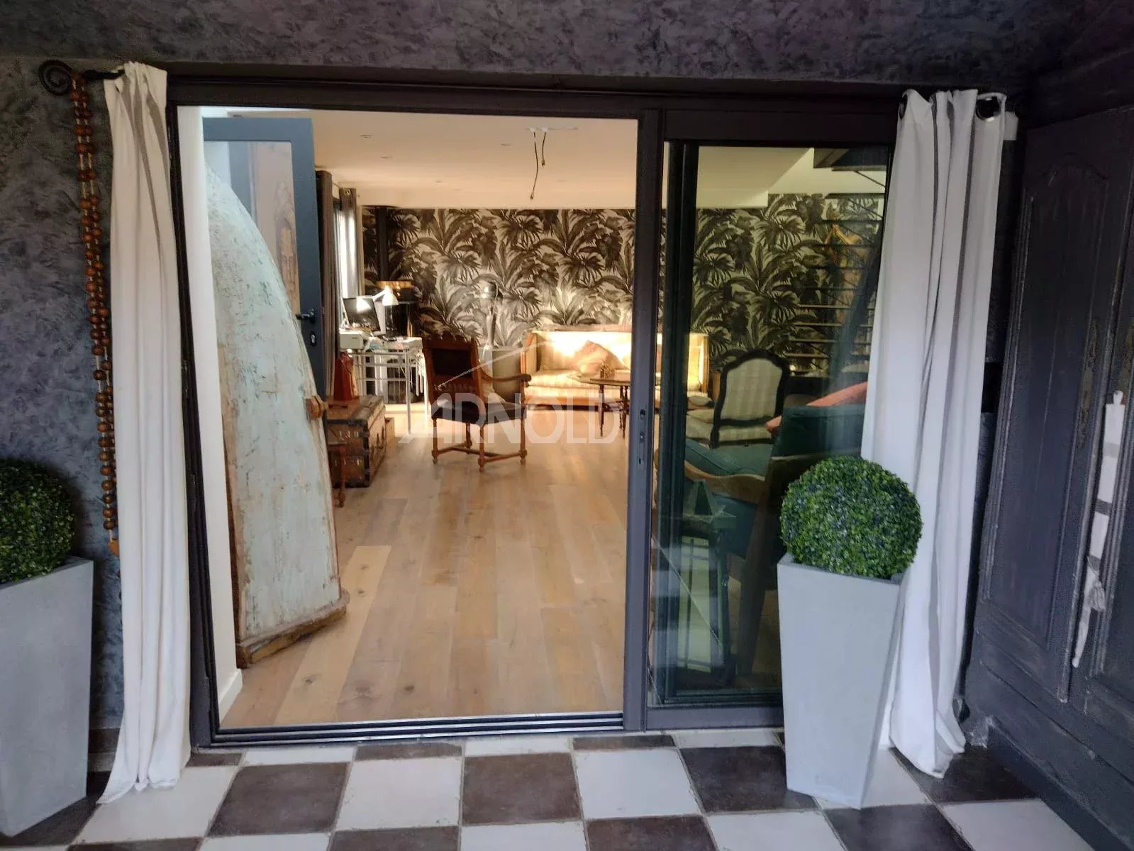 Sliding glass doors open to a stylish living room with palm-pattern wallpaper, wood floor, and vintage furniture including a tall leaning surfboard or panel on the left.