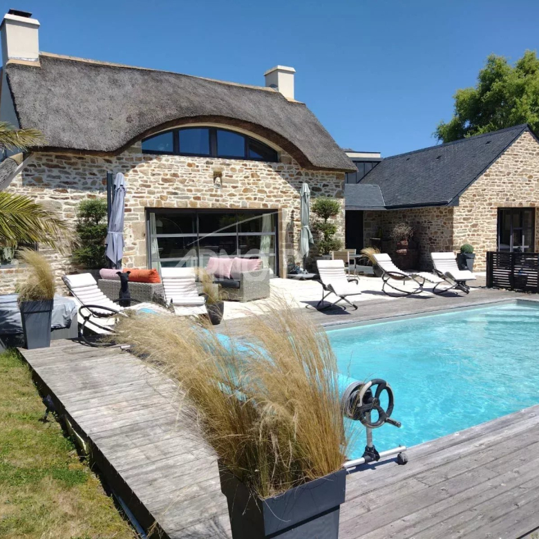 Stone cottage with a thatched roof, arched window, and a rectangular pool on a wooden deck with white sun loungers and seating.