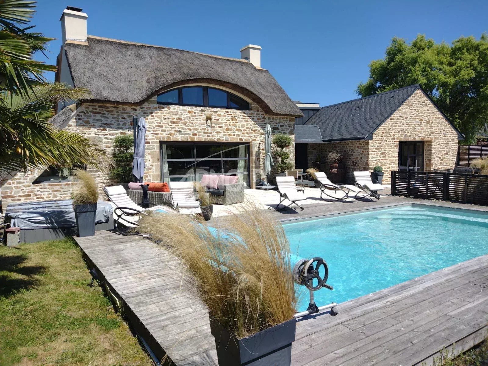 Stone cottage with a thatched roof, arched window, and a rectangular pool on a wooden deck with white sun loungers and seating.