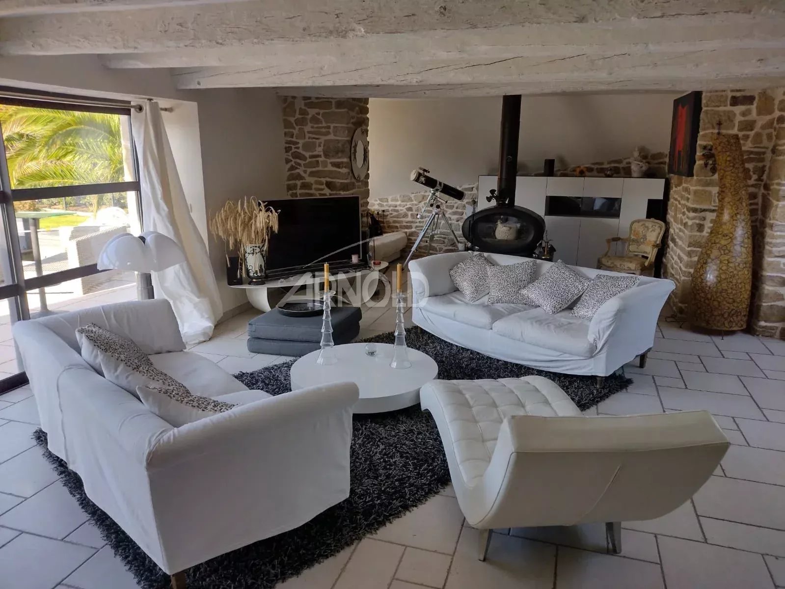 Bright living room with white sofas around a round coffee table on a black shag rug; stone walls and a telescope near a wood-burning stove.