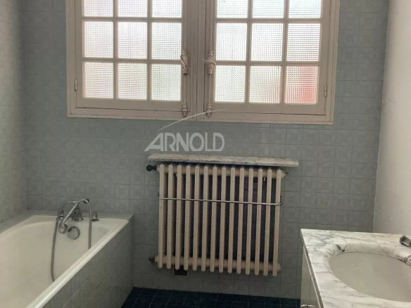 Salle de bain avec baignoire, radiateur et lavabo, murs carrelés en bleu et fenêtres à grille.
