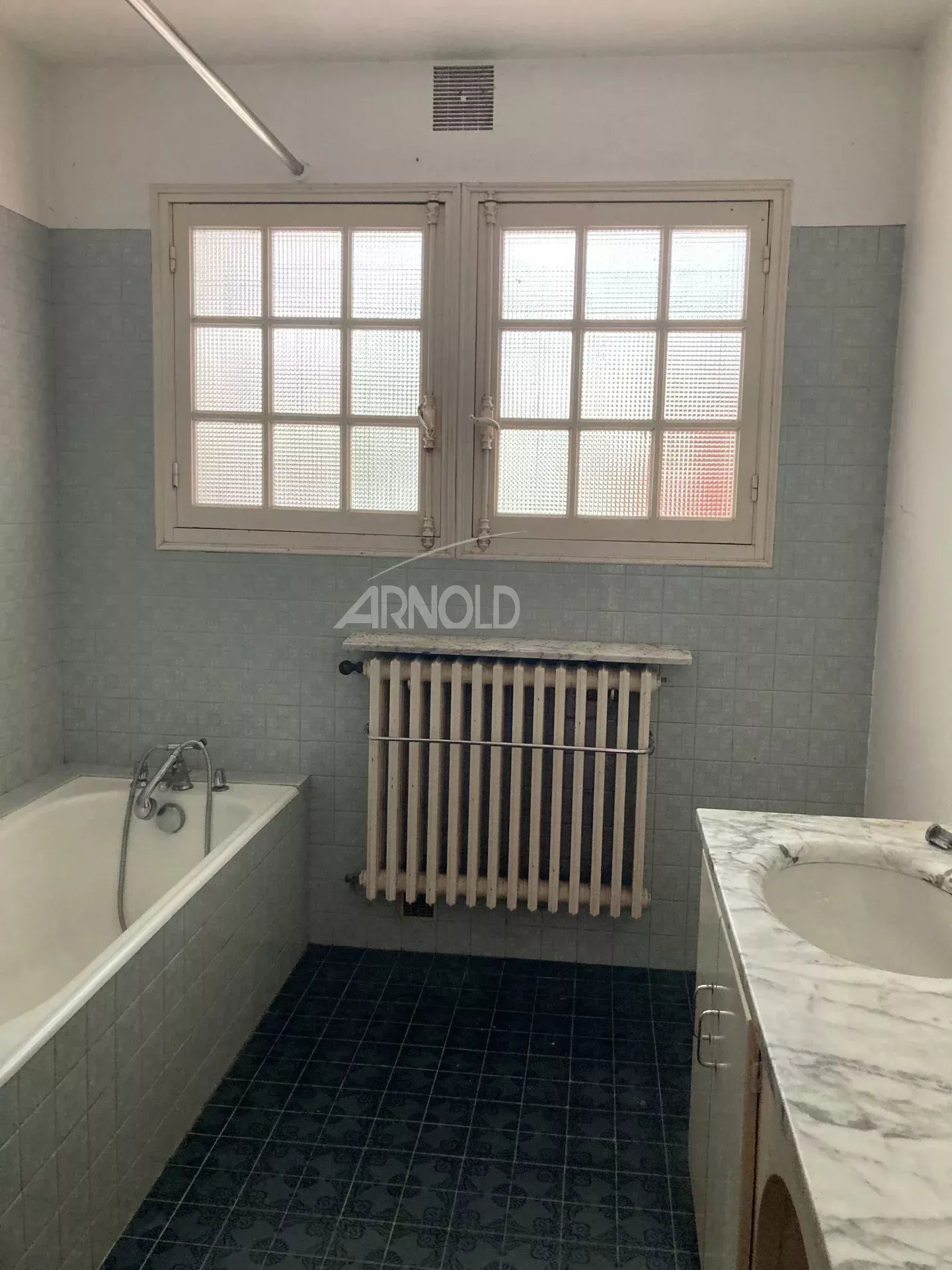 Salle de bain avec baignoire, radiateur et lavabo, murs carrelés en bleu et fenêtres à grille.
