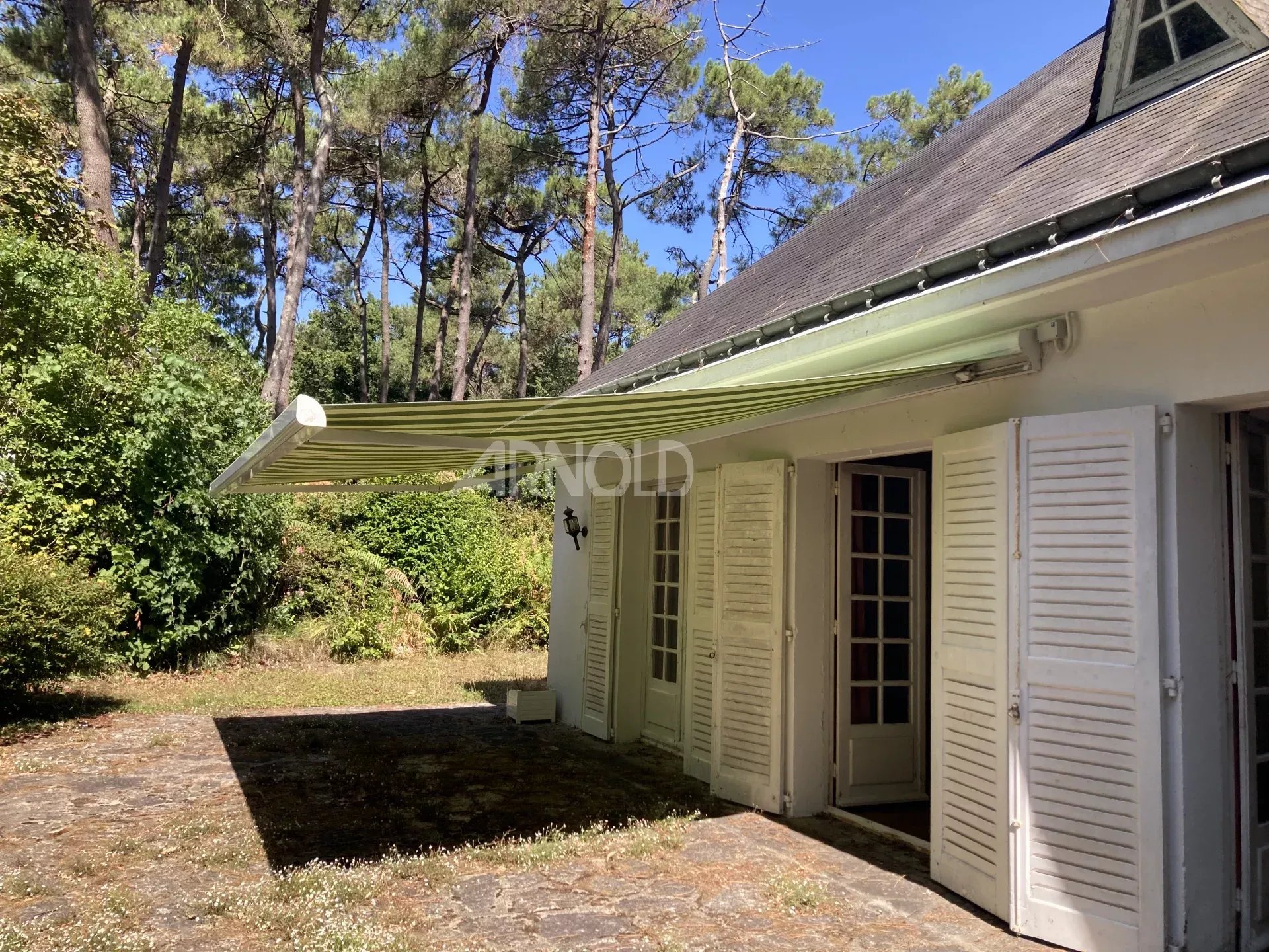 Maison avec auvent, située dans un jardin arboré, entourée d'arbres.