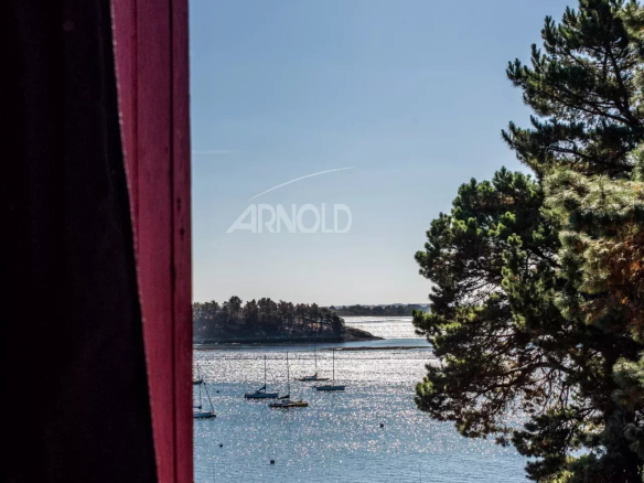 Vue panoramique sur le Golfe du Morbihan depuis une fenêtre, avec des voiliers et des arbres au premier plan.