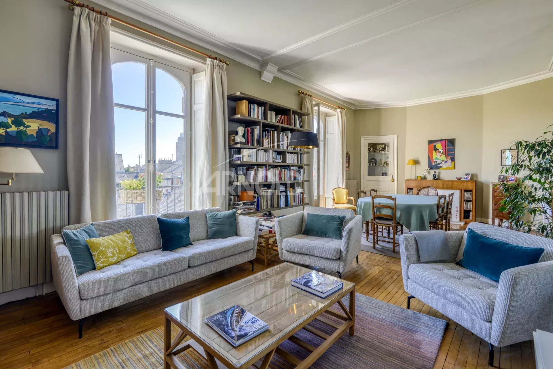 Salon lumineux d'un appartement T6 avec canapés et bibliothèque, fenêtres ouvertes sur l'extérieur.