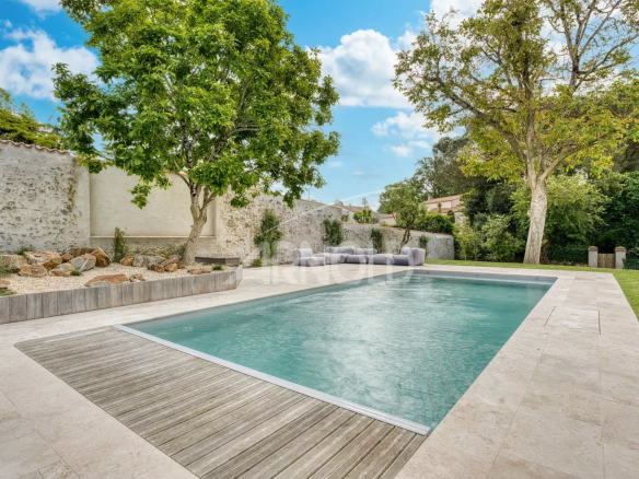 Swimming pool surrounded by a garden with trees and rocks, near an old building.