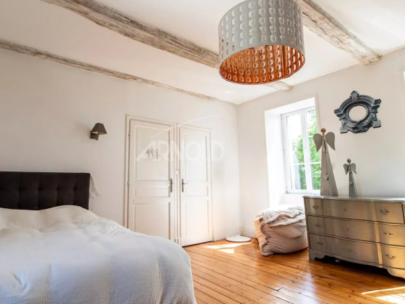 Chambre lumineuse avec lit, commode et fenêtres, poutres apparentes au plafond.
