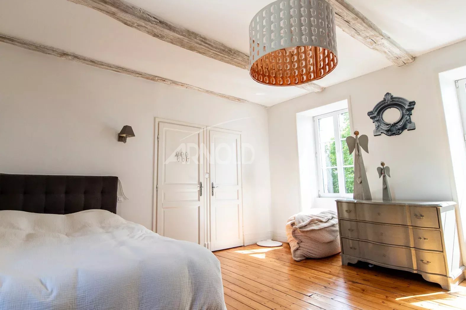 Chambre de charme avec poutres apparentes Chambre lumineuse avec lit, commode et fenêtres, poutres apparentes au plafond.