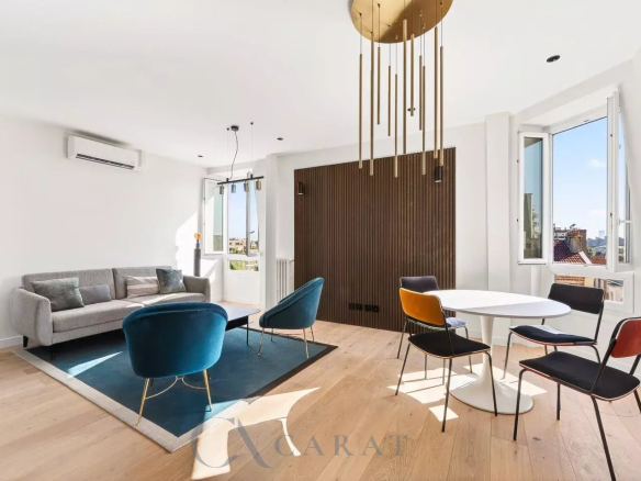Bright open-concept living area with a gray sofa, blue accent chairs, and a round white dining table by large windows.