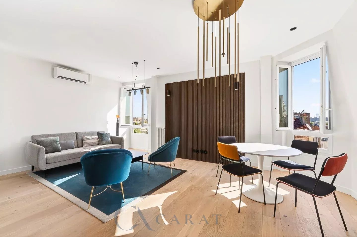 1dd9e41a0727b7c3693413147b5a76fe_2cacbf6ba8_1440-original Bright open-concept living area with a gray sofa, blue accent chairs, and a round white dining table by large windows.