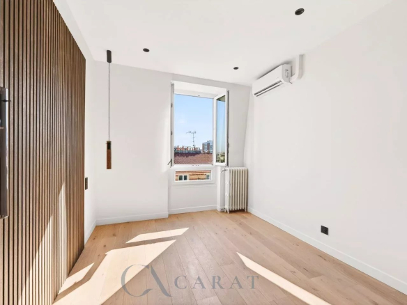 Sunlit empty room with a wood-paneled wall on the left, a hanging pendant light, and a window showing rooftops outside; radiator and AC unit present.