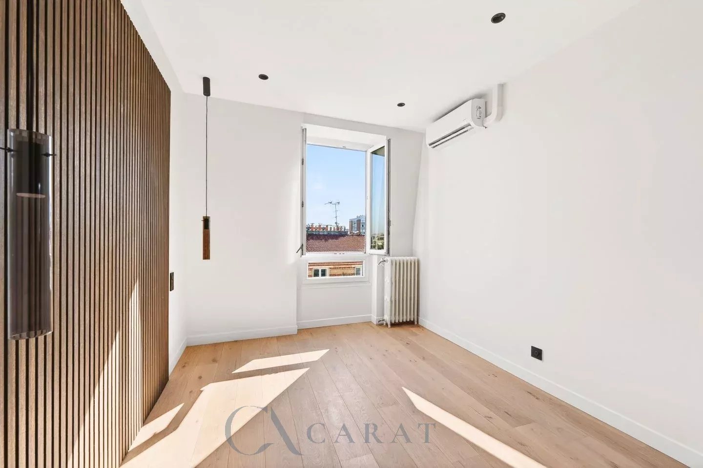 3e85e77a2b3003d5c6fb892de6774015_bf33b4ee3d_1440-original Sunlit empty room with a wood-paneled wall on the left, a hanging pendant light, and a window showing rooftops outside; radiator and AC unit present.