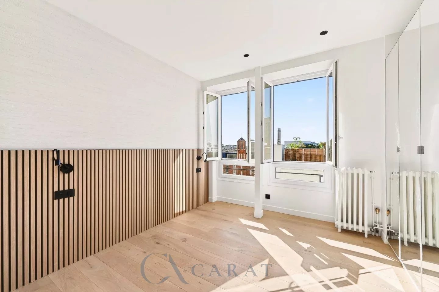 4bfc57a3e9f4a3882eaf95bde8beb081_5274e270e0_1440-original Bright, empty room with large open windows and a city skyline view, light wood floor, and white walls.