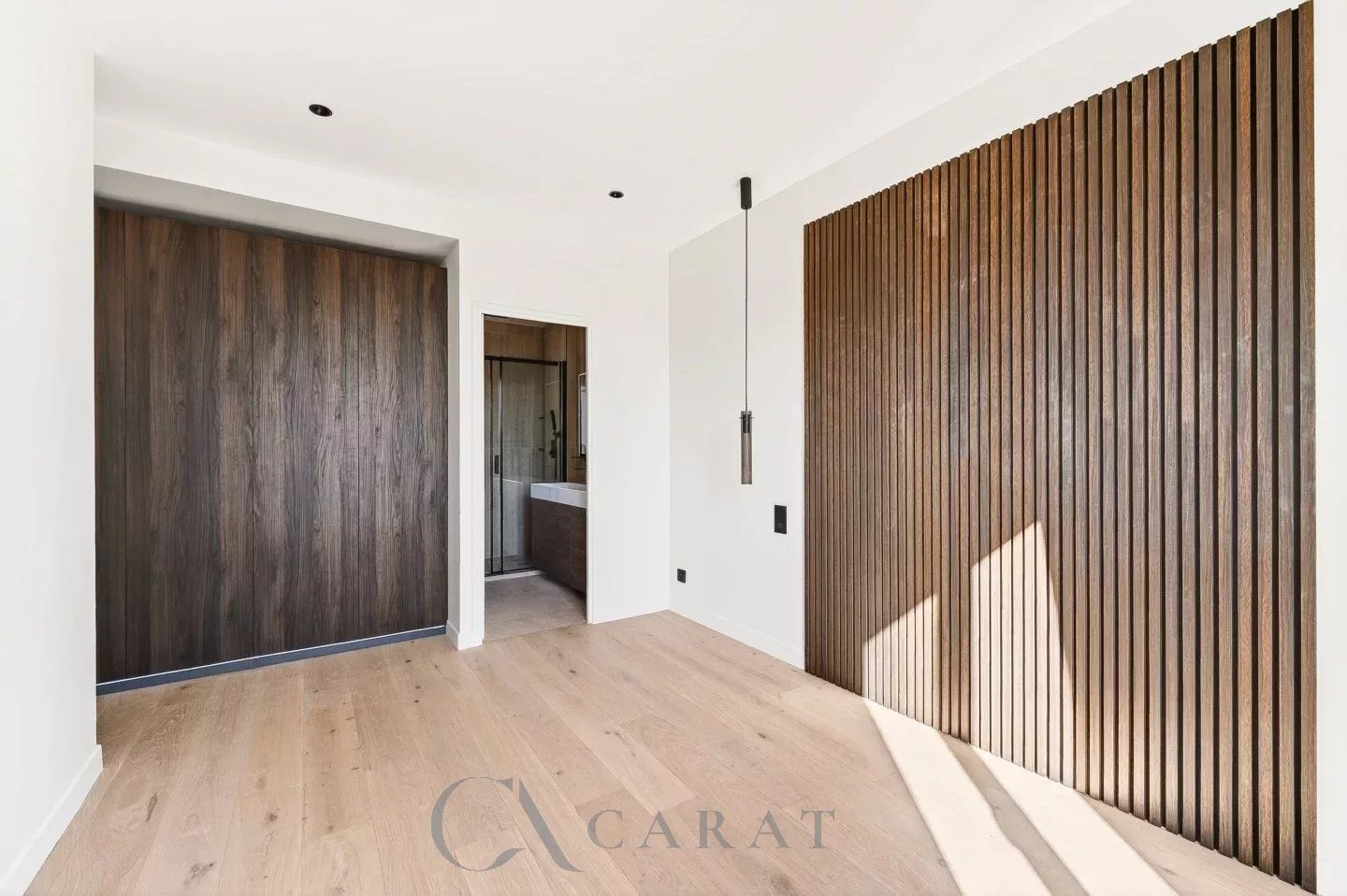 4d8dbd539c0079c9d282cb9256e10abb_f8a23d91a8_1440-original Modern empty room with white walls, light wood floor, a tall dark wood sliding door on the left, and a vertical wood slat wall on the right; doorway to a bathroom in the center.