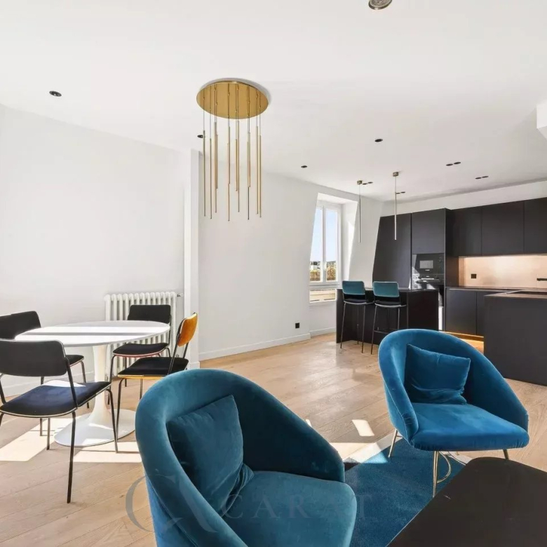 Open-plan living space with blue velvet chairs, a white round dining table, and a dark contemporary kitchen with gold pendant lights.
