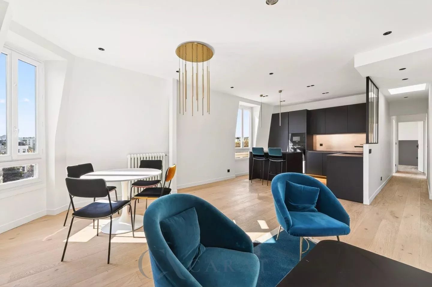 7180d88cfc69da62912049136b716fec_0e1971cb4c_1440-original Open-plan living space with blue velvet chairs, a white round dining table, and a dark contemporary kitchen with gold pendant lights.