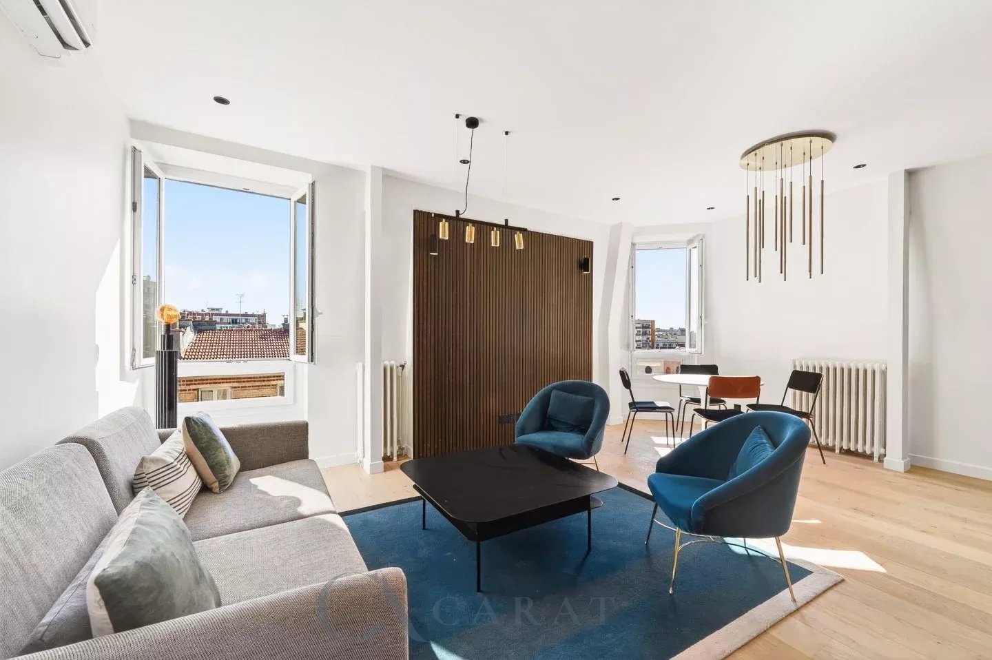 8a6a3c4d8ae8ad9f6846b0d0ced5b49e_0d09c566b4_1440-original Bright living room with a gray sofa, blue accent chairs, and a wooden slat divider, under sunny windows on the left and right side.