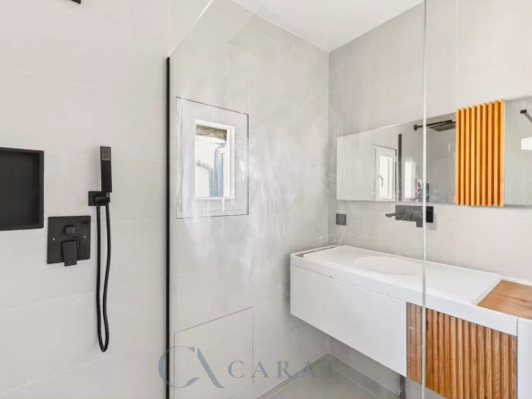 Modern grey-tiled bathroom with a glass shower partition, black fixtures, and a white vanity with wood accents.
