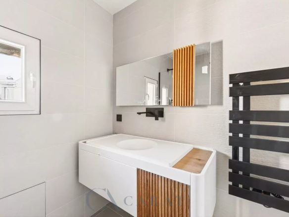 Modern bathroom with white vanity featuring wood slat panel, large mirror, and black wall-mounted faucet and radiator letters; window on left wall.