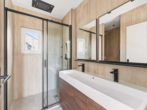 Modern bathroom with tan tile walls, a double white sink vanity with black fixtures, large mirrors, and a glass shower enclosure.
