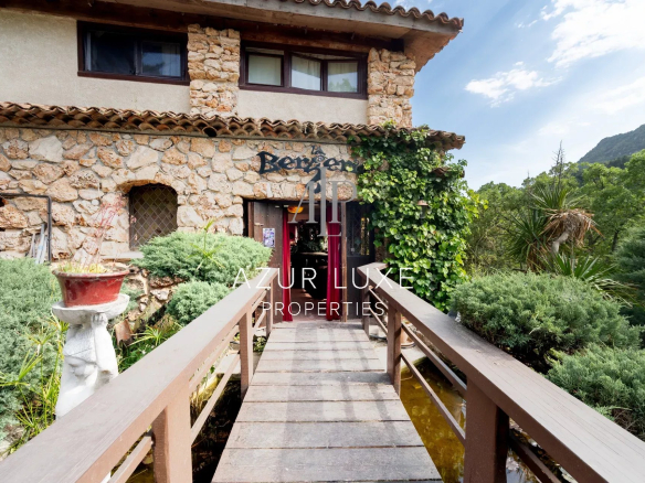 Entrance to an old stone hotel-restaurant with a wooden bridge, surrounded by greenery in Castillon.