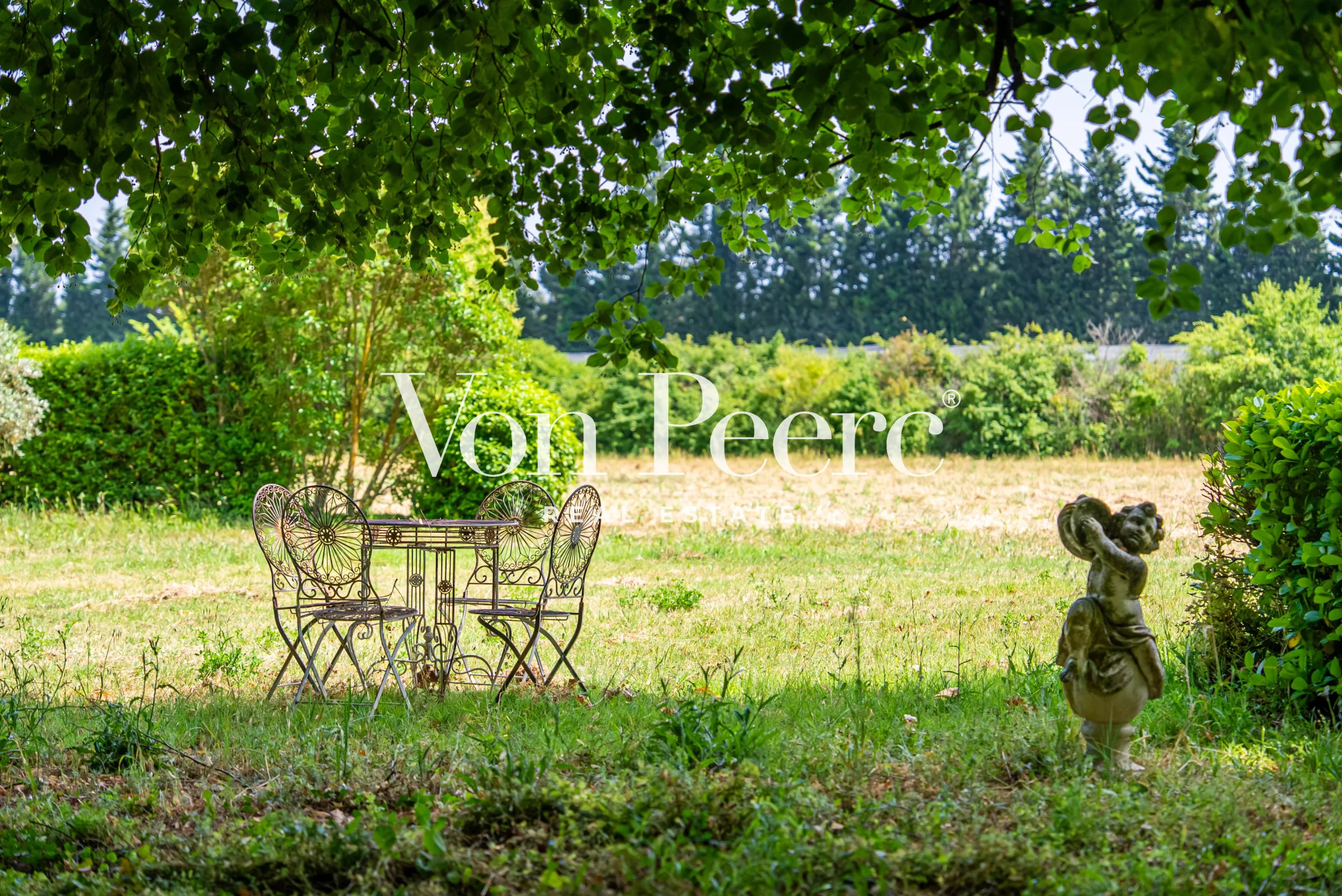 Table et chaises en fer forgé dans un jardin verdoyant avec des arbres en arrière-plan.