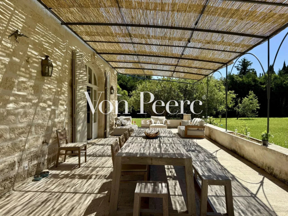 Terrasse d'une maison ancienne rénovée avec mobilier en bois, ombragée par un auvent, jardin verdoyant en arrière-plan.