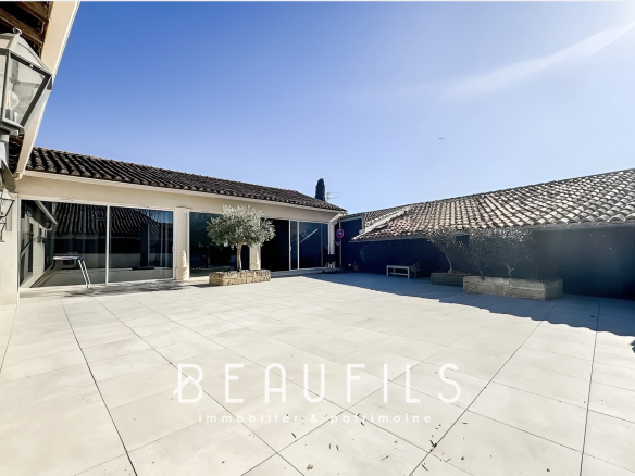 Sunny courtyard of a house with a large tiled patio, glass sliding doors, and potted olive trees under a blue sky.