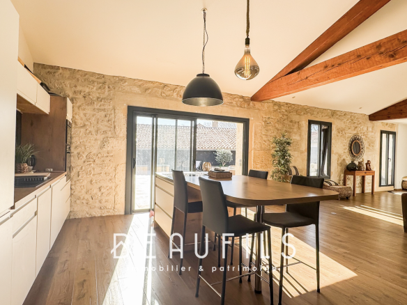 Open-plan kitchen and dining area with a large wooden table, black chairs, stone walls, and exposed wooden ceiling beams. Sunlight streams through glass doors to the outside.