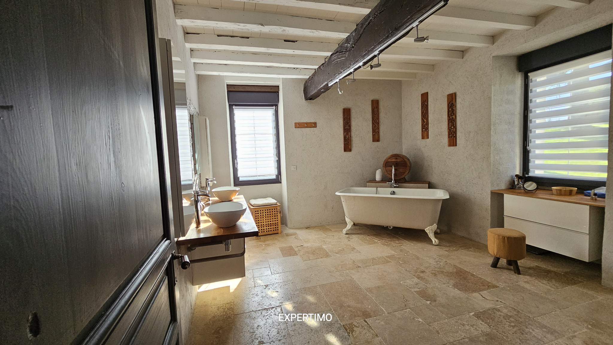 Rustic bathroom with a freestanding clawfoot tub, wooden beams, and a double sink vanity under large windows.