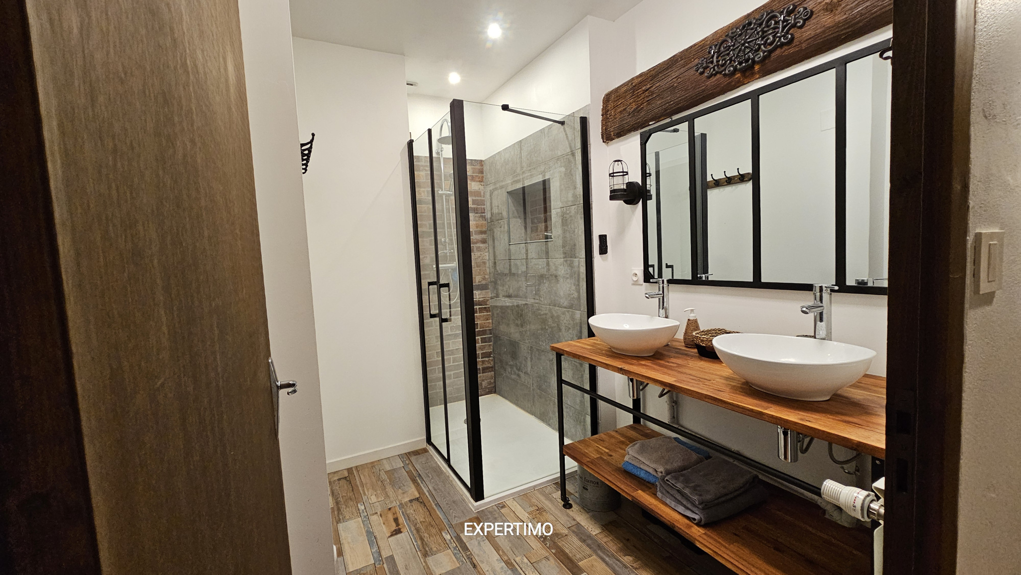 Modern rustic bathroom with double sinks on a wooden counter, large mirror, and glass-enclosed shower.