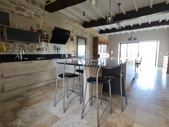 Open-concept kitchen with stone walls, dark countertops, and a glass-top island surrounded by four metal-bar stools. The space has exposed wooden beams and a bright dining area in the background.