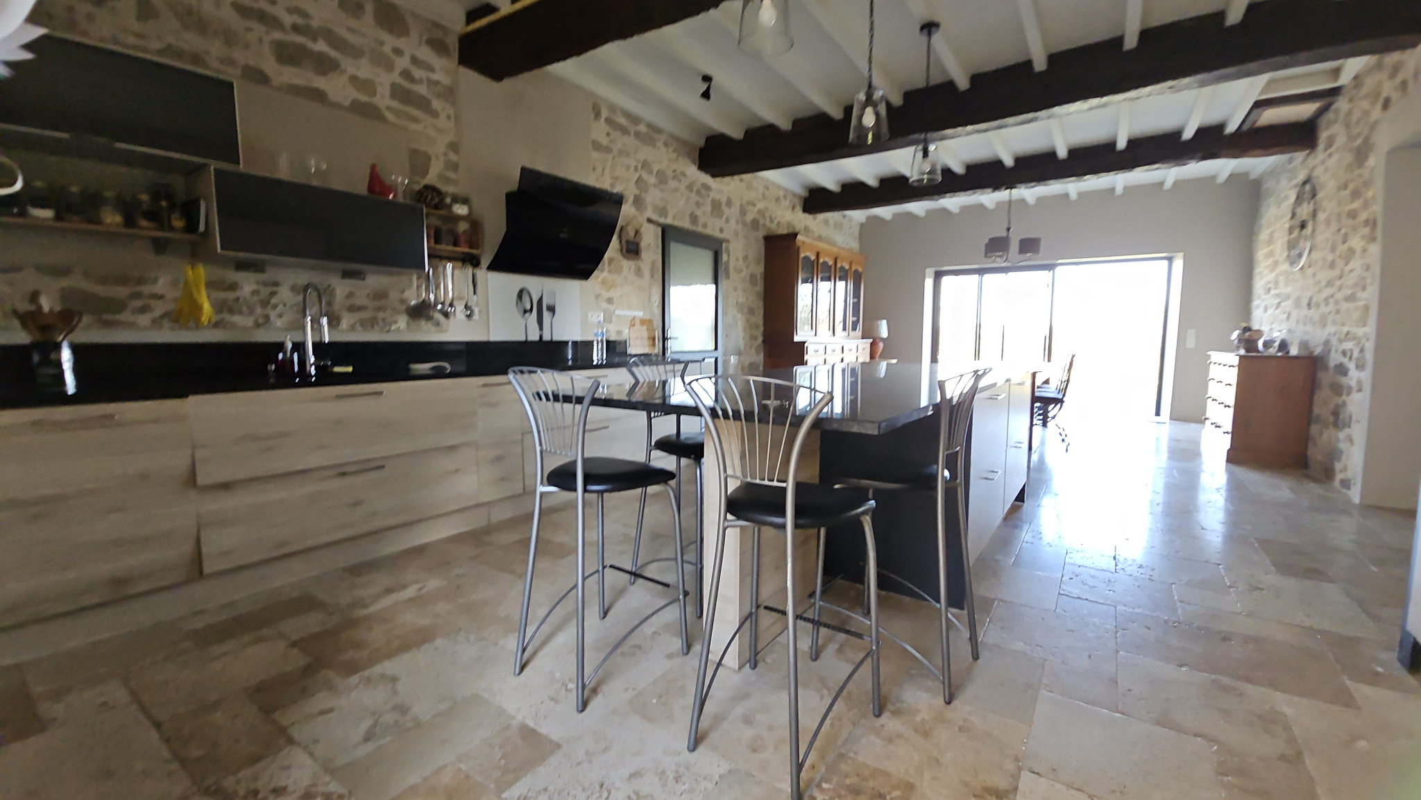 Open-concept kitchen with stone walls, dark countertops, and a glass-top island surrounded by four metal-bar stools. The space has exposed wooden beams and a bright dining area in the background.