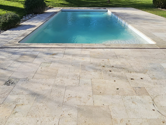 Rectangular backyard pool with beige stone patio, green lawn, and trees under a blue sky.