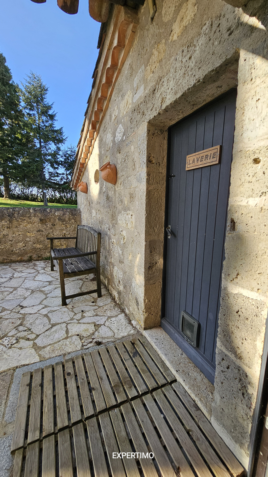Blue door labeled 'LAVERIE' set in a rustic stone wall, with a wooden bench and stone courtyard in bright sunlight
