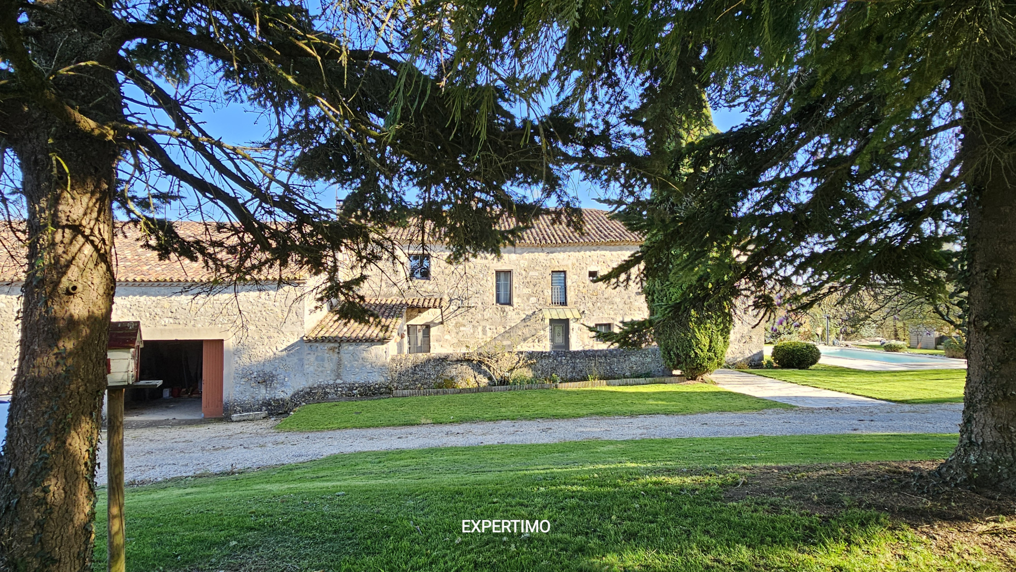 Stone farmhouse with a red-tiled roof framed by overhanging evergreen branches, with a gravel path and manicured lawn in the foreground.”]