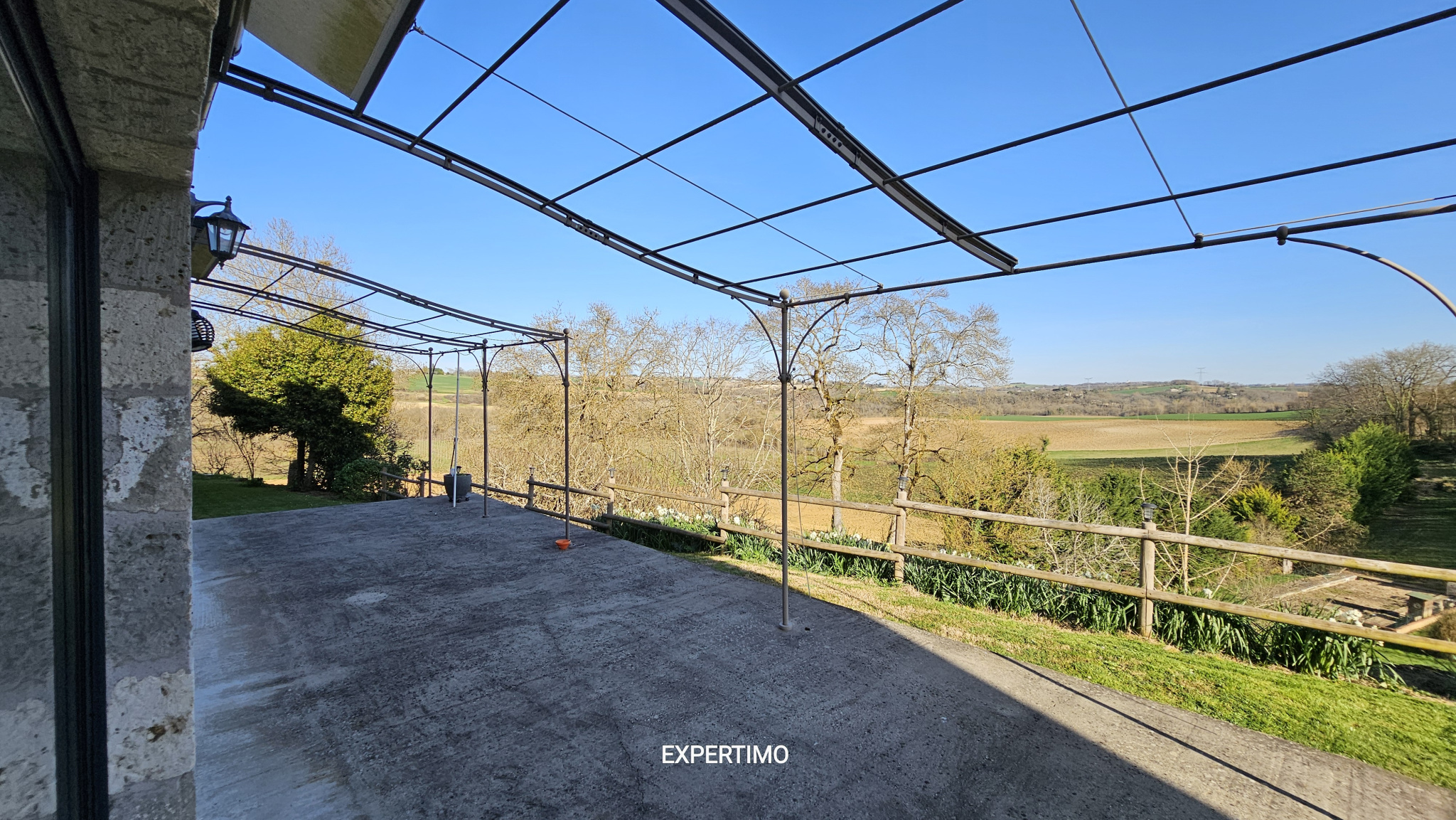 Outdoor concrete patio under a metal pergola frame with a scenic countryside view and a wooden railing beyond; clear blue sky.