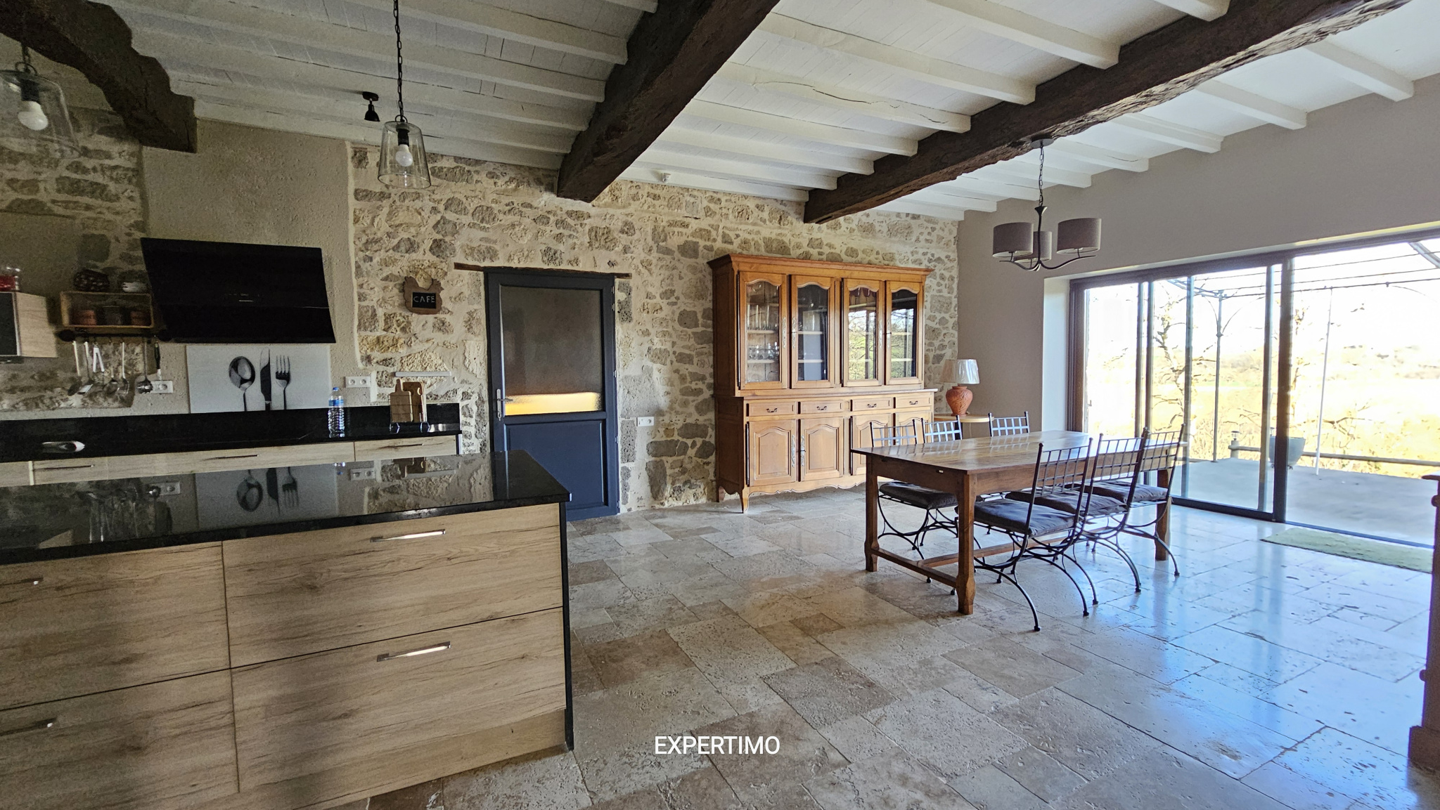 Rustic kitchen-dining area with exposed stone wall, wooden beams, and large glass doors opening to an outdoor view.