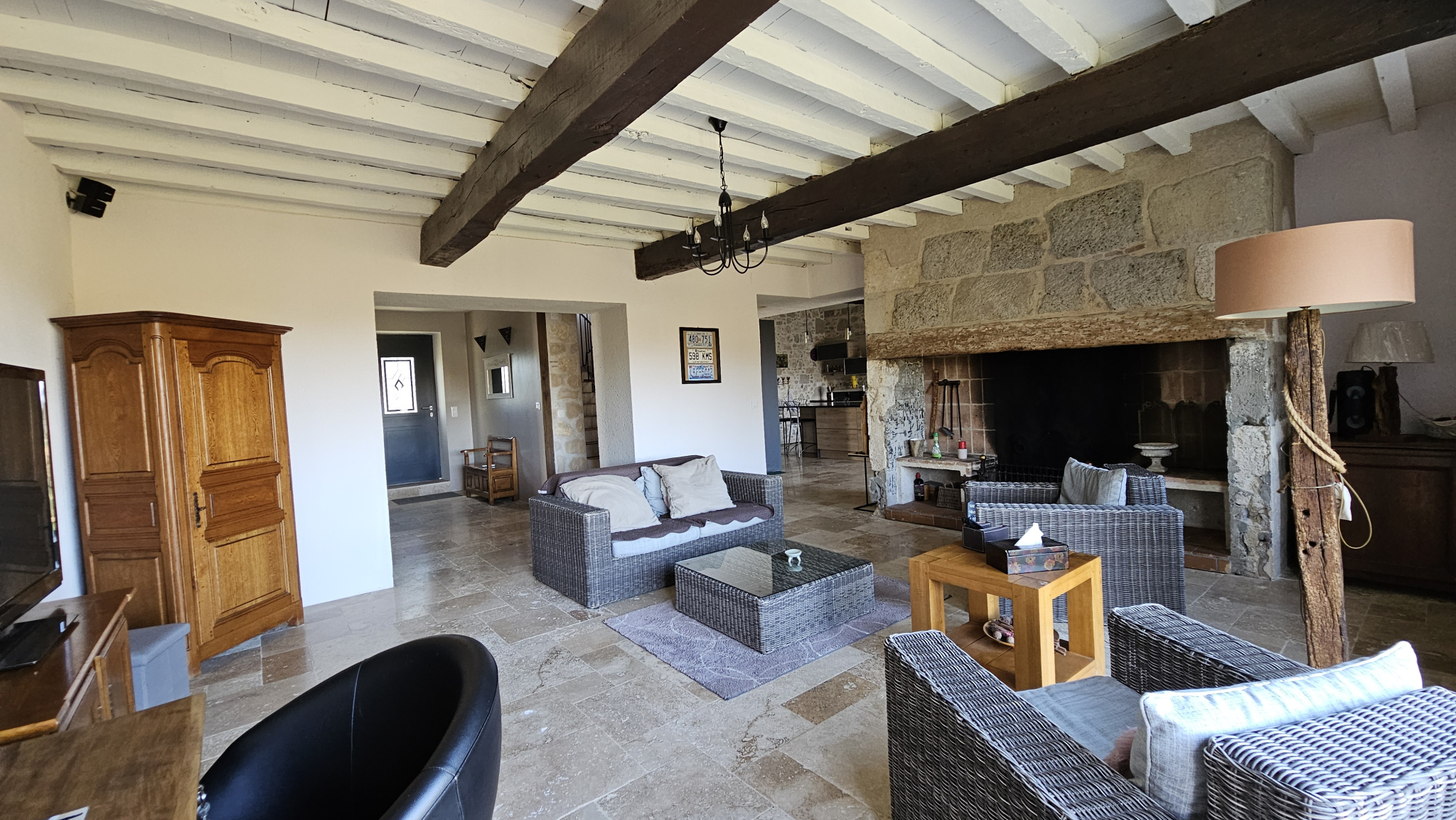 Rustic living room with a large stone fireplace, whitewashed ceiling beams, and wicker seating around a glass coffee table.