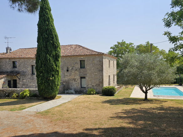 Stone house with red-tiled roof and a tall cypress in the front yard, pool to the right on a sunny day.