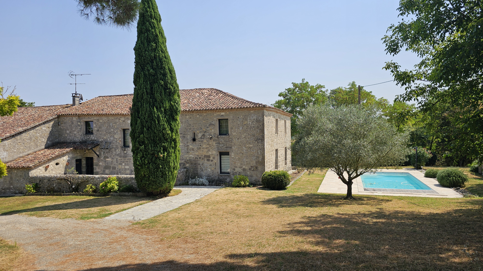 Stone house with red-tiled roof and a tall cypress in the front yard, pool to the right on a sunny day.