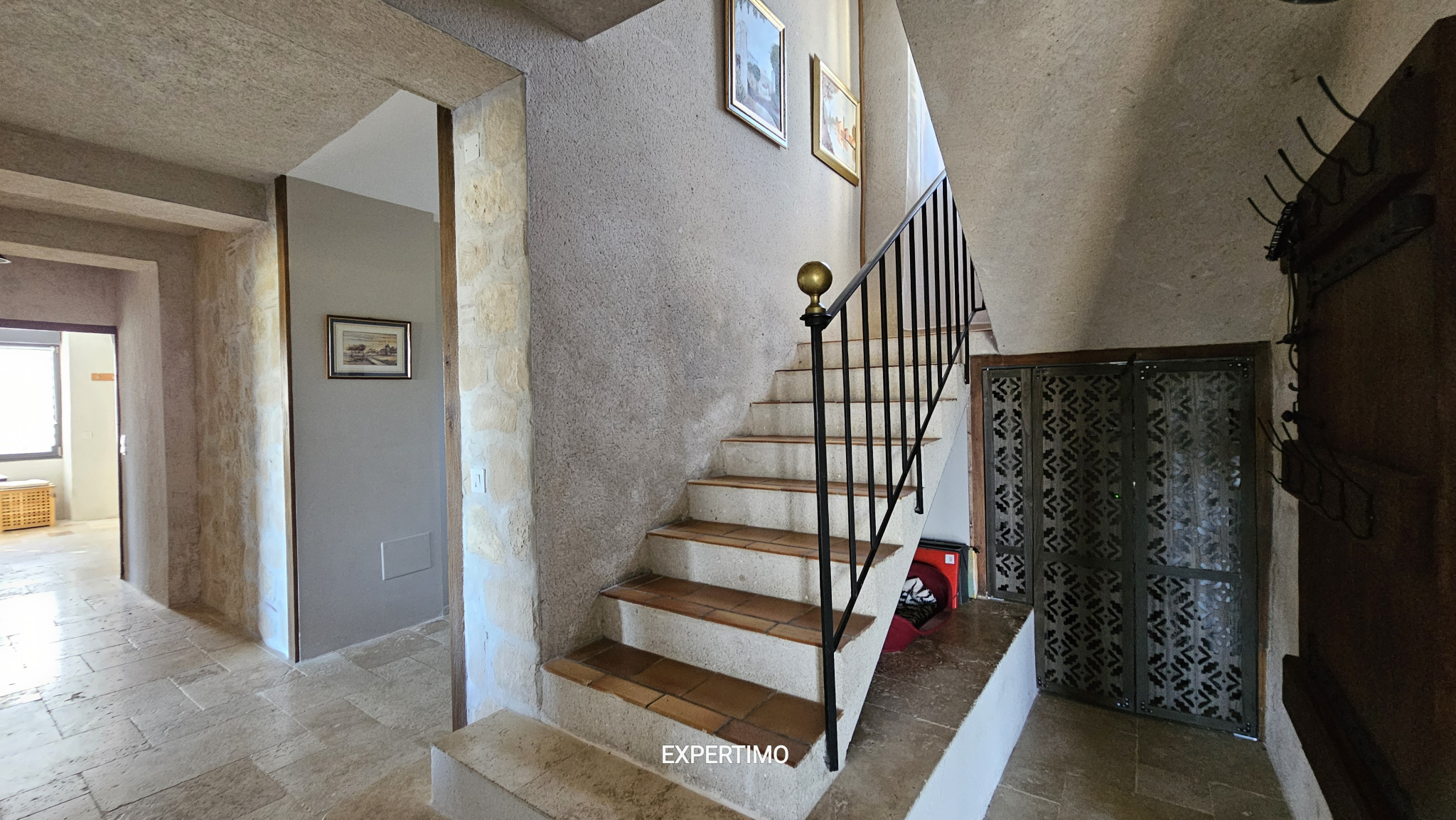 Stone-walled interior with a concrete staircase and black metal railing, and framed paintings on the wall above the stairs.